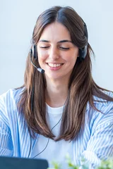 Smiling customer service representative wearing a headset and working on a laptop, offering friendly remote support in a modern office setting and engaging in a voice or video call. © simona