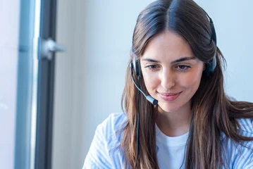 Smiling customer service representative wearing a headset working remotely at a laptop, providing friendly business support and communication in a bright modern office © simona