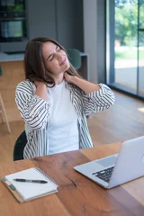 Young woman rubbing her neck in discomfort while working on a laptop at a home office desk, showing neck pain, fatigue and the need for ergonomic breaks and posture correction. © simona