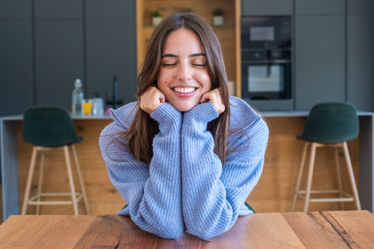 Smiling young woman in a cozy blue sweater leans on a kitchen table with eyes closed, radiating relaxed, happy contentment in a modern home setting.