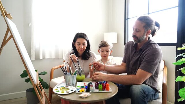 The family&mdash;mother, father, and son&mdash;are learning to paint with watercolors. The mother is teaching, the parents are teaching their son, and they are chatting as a hobby in the living room at home.