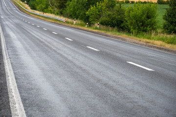Obraz premium An empty asphalt highway with dashed white dividing lines gently curves through a rural landscape bordered by lush green trees