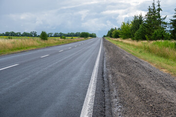 Obraz premium A straight, empty asphalt highway stretches towards the horizon through a rural landscape under a cloudy, overcast sky