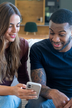 Two friends, a woman and a man, smile and look at a smartphone together on a couch at home, sharing a moment of surprise and connection while casually using technology.