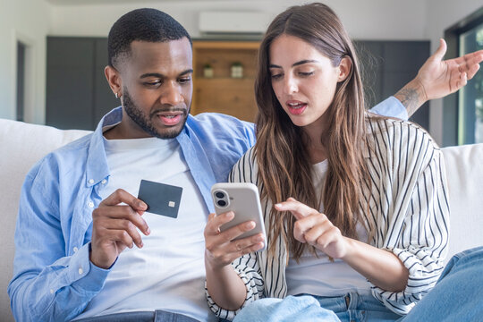 Young mixed-race couple shopping online at home using a smartphone and credit card while sitting together on a sofa, planning purchases
