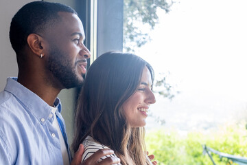 Fototapeta premium Couple standing by a window, looking out together and smiling lovingly as they enjoy a peaceful moment and bright view, showing affection, connection and optimism.