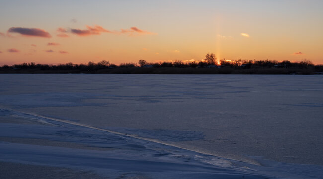 Beautiful winter sunset over a frozen river with warm golden reflections on the icy surface. Peaceful seasonal nature landscape during cold weather.