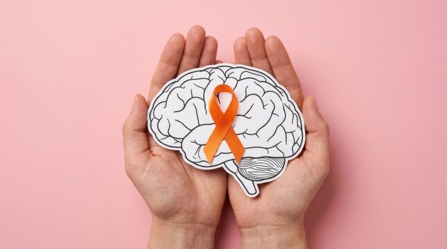 Top view of hands holding a human brain paper cutout with an orange awareness ribbon for Multiple Sclerosis, ADHD, and Kidney Cancer awareness on pink background