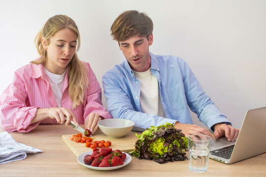 Young couple preparing a healthy salad together at home while using a laptop for a recipe, chopping vegetables and strawberries on a wooden board, sharing a relaxed cooking and learning moment.