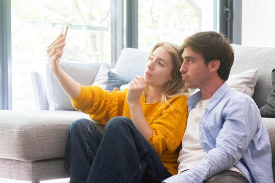 Young couple sitting on a living room floor taking a playful selfie together, relaxed and cozy at home, smiling and making faces while leaning against a sofa in natural daylight.
