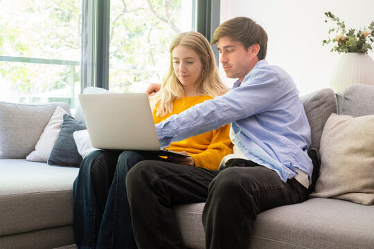 Couple sitting on a couch at home using a laptop together, browsing and planning while relaxed, sharing ideas and shopping online in a bright living room