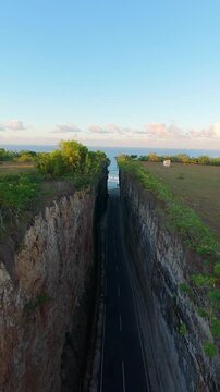 Vertical video. Smooth Cinematic Aerial Flight Above Deep Road Cut Through Coastal Cliffs At Pantai Tanah Barak, Wide Angle Sunrise View Toward Ocean Horizon And Green Landscape, Journey And Discovery