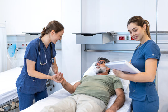 Two female doctors checking on older male patient in hospital bed after successful operation. Friendly physician holding his hand. Healthcare, medicine and postoperative clinical recovery concept.