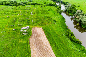 Obraz premium Aerial view of overgrown abandoned farmland with decaying greenhouse ruins, remnants of old agricultural structures set in a vibrant green meadow beside a calm winding river