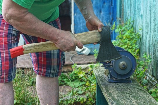 A man holding and sharpens an ax iron blade on an electric sharpening machine. Close-up of the process of sharpening a tool with a wooden handle in the courtyard of the workshop.