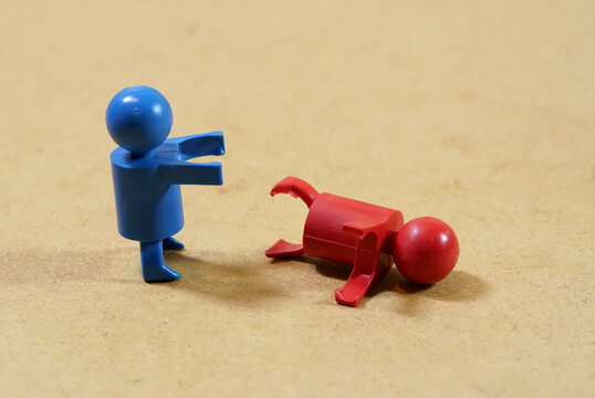 Blue toy figure standing with arms extended beside a fallen red figure on a brown surface, symbolic scene of victory, defeat, conflict, dominance, assistance, rescue or offering help, with copy space