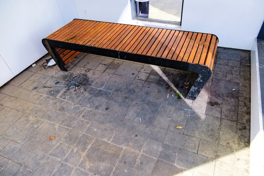 A neglected urban bench sits on a stained, grimy tiled floor, surrounded by wet puddles, scattered trash, and debris, with a sharp shadow suggesting an unkempt public area