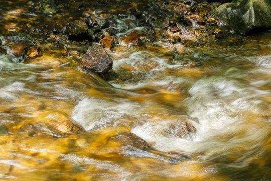 Gold colored water stream flowing over rocks and smooth stones in a river with motion blur effect, creating a sense of movement and tranquility.