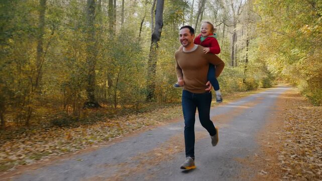 man carrying child running on forest trail with autumn leaves and dirt path, joyful family activities, parent-child bonding, side view, slightly low angle, vibrant natural setting.