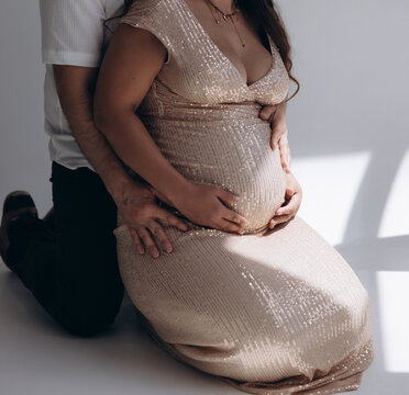 Side view of pregnant woman in sequin dress sitting on floor with husband hugging her belly