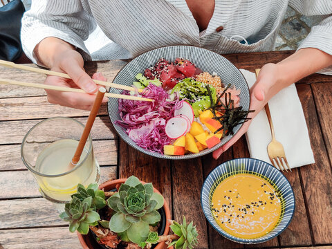 Woman eating tasty colorful healthy natural organic vegetarian Hawaiian poke bowl using asian chopsticks on rustic wooden table. Healthy natural organic eating concept.