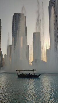 Traditional boat with people cruising on water in front of modern city skyscrapers and fountains in Dubai. 