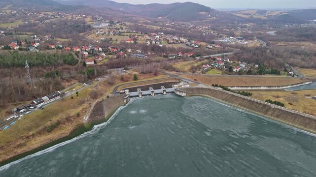 In Świnna Poręba, the Zapora Świnna Poręba dam spans the frozen surface of Lake Mucharskie amid surrounding hills and forest, captured in an aerial view of the winter landscape.