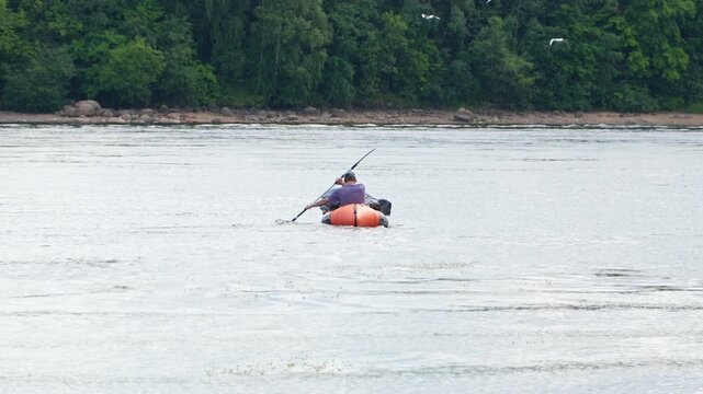 Man paddling in packraft or inflatable kayak on a wide river with green forest background. Solitary traveler paddling on a river during water tourism trip concept of freedom and adventure.