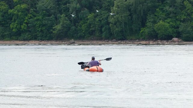 Man paddling in packraft or inflatable kayak on a wide river with green forest background. Solitary traveler paddling on a river during water tourism trip concept of freedom and adventure.