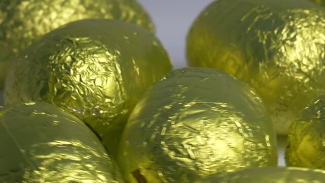 Close-up of chocolate Easter eggs wrapped in shiny golden foil rotating on a white plate, highlighting their textured metallic surface under bright lighting.