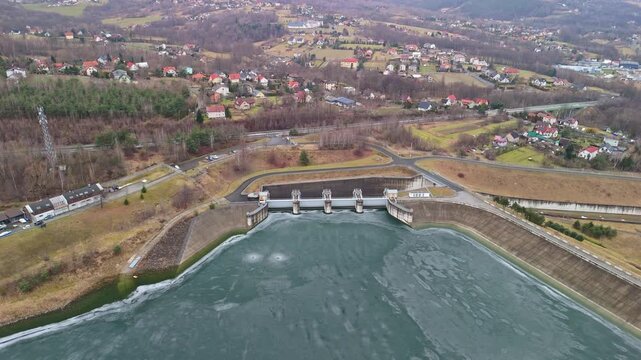 In Lesser Poland, the Zapora Świnna Poręba dam borders the frozen Jezioro Mucharskie within a rural valley landscape, with its concrete structure and spillway viewed from a drone.