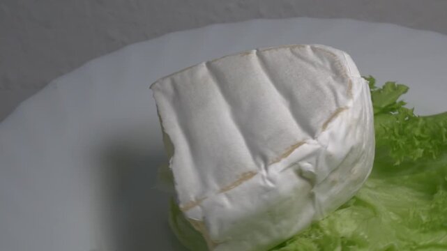 Close-up of a piece of Brie cheese on fresh green lettuce leaves rotating on a white plate, showcasing its soft texture and white rind under studio lighting.