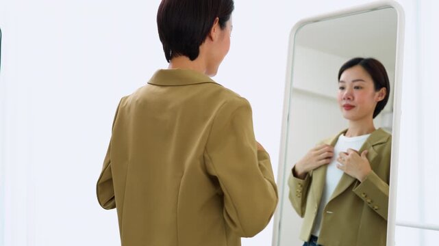 Confident Asian female getting dressed in front of a mirror, preparing for a workday or job interview. Concept of elegant wardrobe selection and modern lifestyle.