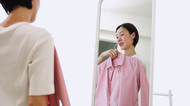 Cheerful Asian female holding hangers with casual shirts, preparing her outfit in a mirror. Concept of personal style development, dressing up and modern lifestyle.