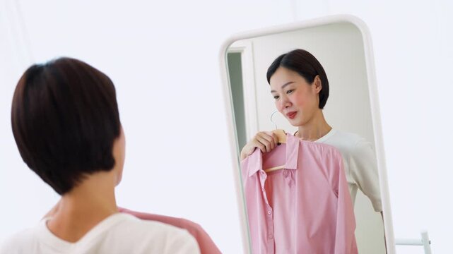 Cheerful Asian female holding hangers with casual shirts, preparing her outfit in a mirror. Concept of personal style development, dressing up and modern lifestyle.