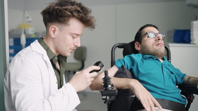 Young specialist in a lab coat helping a male patient with spina bifida by adjusting the joystick controls on his motorized wheelchair