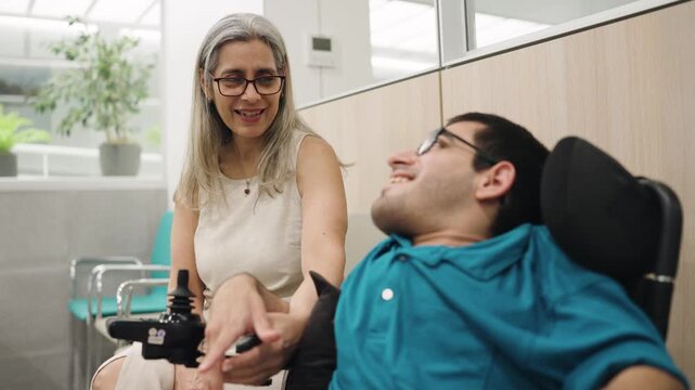 Caring mother sitting next to her disabled son in a wheelchair. The young man with spina bifida is smiling and talking with her in the hospital