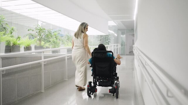 Devoted mother carefully pushing her daughter with spina bifida in a wheelchair. They are moving down a bright, accessible hospital hallway