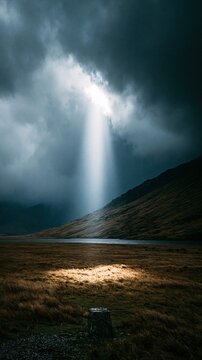 Dramatic single beam of sunlight breaking through dark storm clouds illuminates small patch of dry grass next to calm lake and rolling hills creates moody natural landscape