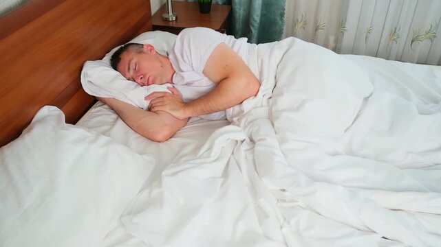 Young man sleeping peacefully in bed under white blanket wooden headboard and in background