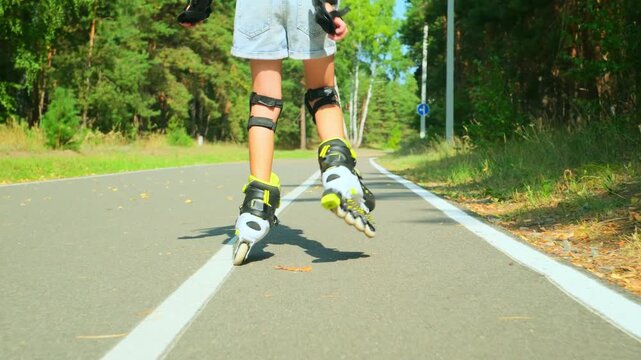 Child girl in casual clothes roller skating on road in forest on sunny summer day.