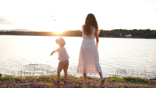 Back view of a mother and her son enjoying a summer evening. A young boy wearing a hat throws stones into the water at sunset