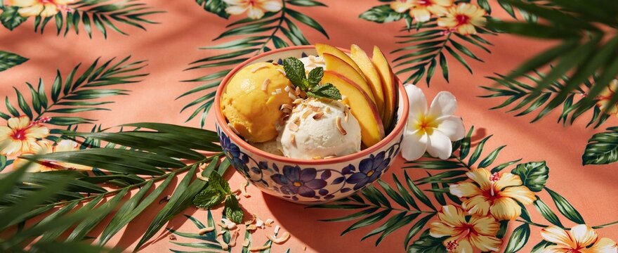Exotic tropical fruit mango coconut ice cream scoops in floral patterned bowl with fresh peach slices mint leaves and frangipani flower on vibrant summer fabric print background sunny day view.