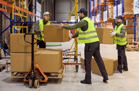 Workers in warehouse loading boxes on pallet. Loaders prepare shipment with pallet jack and stack parcels. Safety vests and logistics ensure fast order flow. Concept of teamwork and logistics.