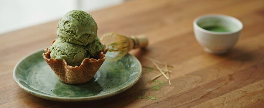 Two scoops of delicious matcha ice cream, presented in a crisp waffle bowl, placed on a green ceramic plate on a warm wooden table with a traditional whisk.