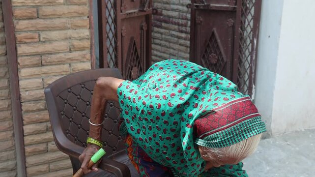 Video of a very elderly Indian woman bending forward while sitting on a chair outdoors. Authentic footage capturing daily life, aging, resilience, and rural Indian culture.
