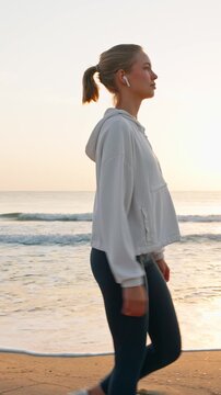 Young fit woman walking fast on sandy beach at sunrise vertical smartphone video orientation female athlete in wireless earphones exercising near sea waves morning workout healthy lifestyle.