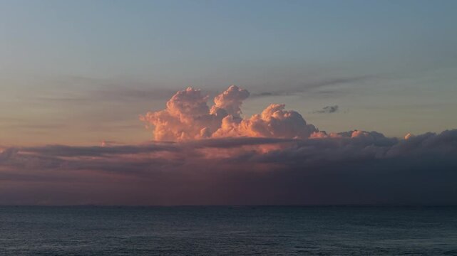 Massive glowing cumulonimbus clouds rising above a calm ocean horizon during sunset. Soft golden light, pastel sky, and tranquil seascape create a dramatic natural landscape perfect for travel, weathe