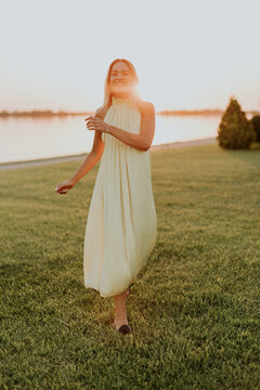 Woman walks on grass by the river during sunset wearing a light colored dress
