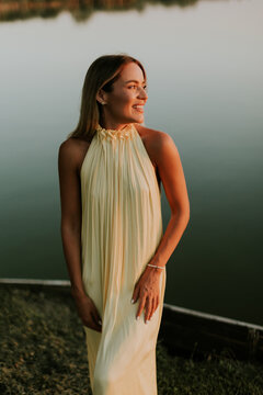 Woman stands by calm water, smiling at sunset near a lake in warm light